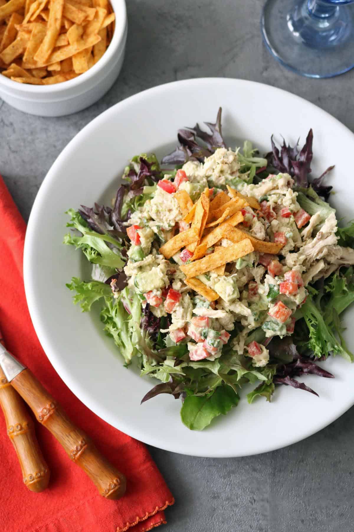 A white plate of Spicy Chicken Salad with Chipotle and Avocado, featuring diced red bell peppers and creamy dressing on leafy greens, topped with crispy tortilla strips. Extra strips and bamboo-handled cutlery sit nearby on a gray surface with a red napkin.