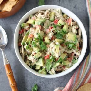 A white bowl filled with Spicy Chicken Salad with Chipotle and Avocado, mixed with diced red bell pepper and cilantro, sits on a gray surface next to tortilla chips, a striped napkin, and a spoon.