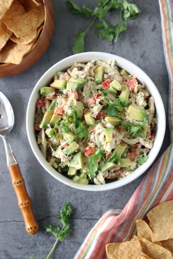 A white bowl filled with Spicy Chicken Salad with Chipotle and Avocado, mixed with diced red bell pepper and cilantro, sits on a gray surface next to tortilla chips, a striped napkin, and a spoon.