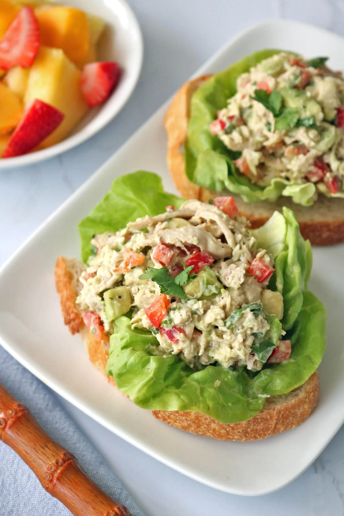 Two slices of bread topped with lettuce and Spicy Chicken Salad with Chipotle and Avocado, served on a white plate. A bowl of mixed fresh fruit sits in the background.