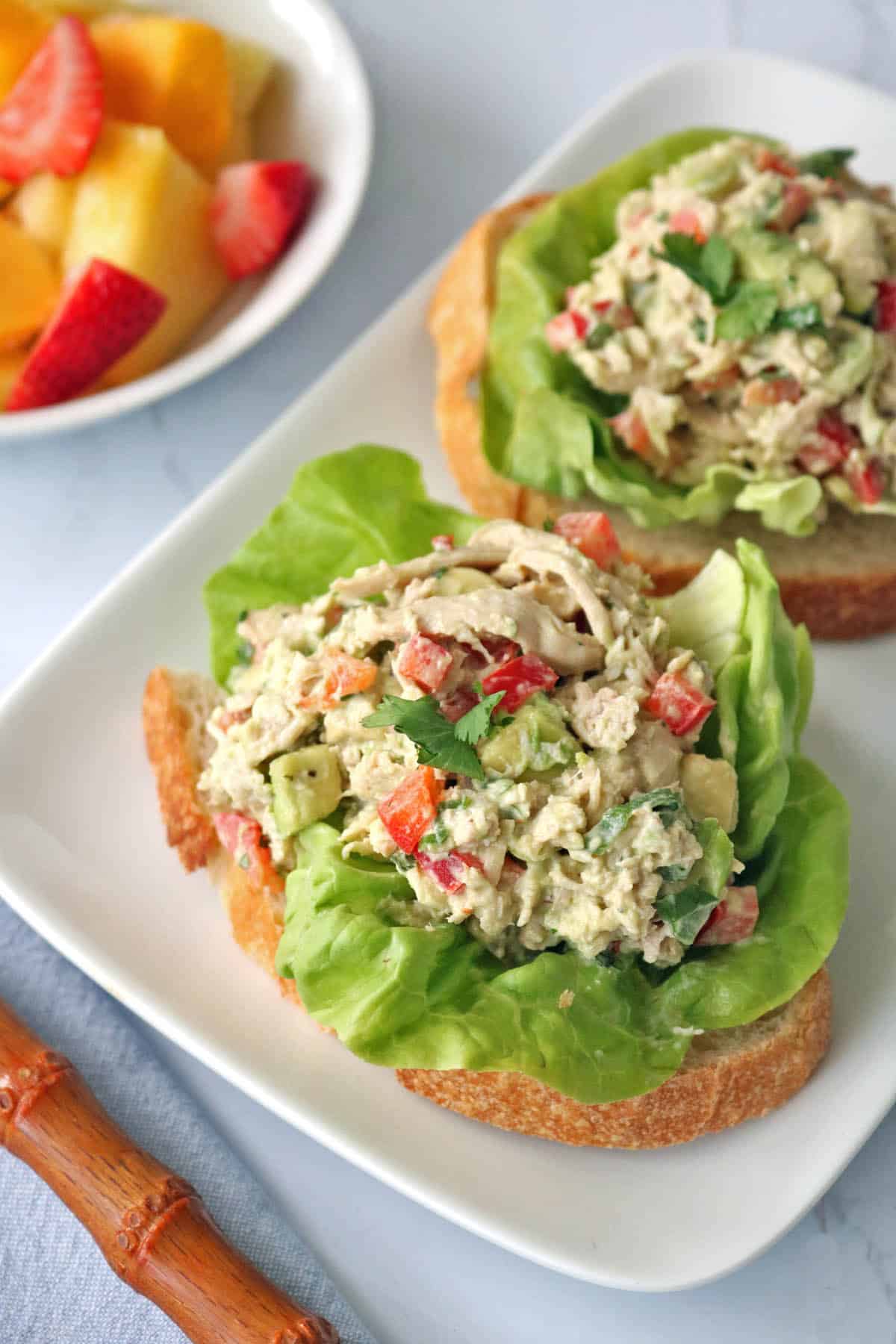 Two slices of bread topped with lettuce and Spicy Chicken Salad with Chipotle and Avocado, served on a white plate. A bowl of mixed fresh fruit sits in the background.