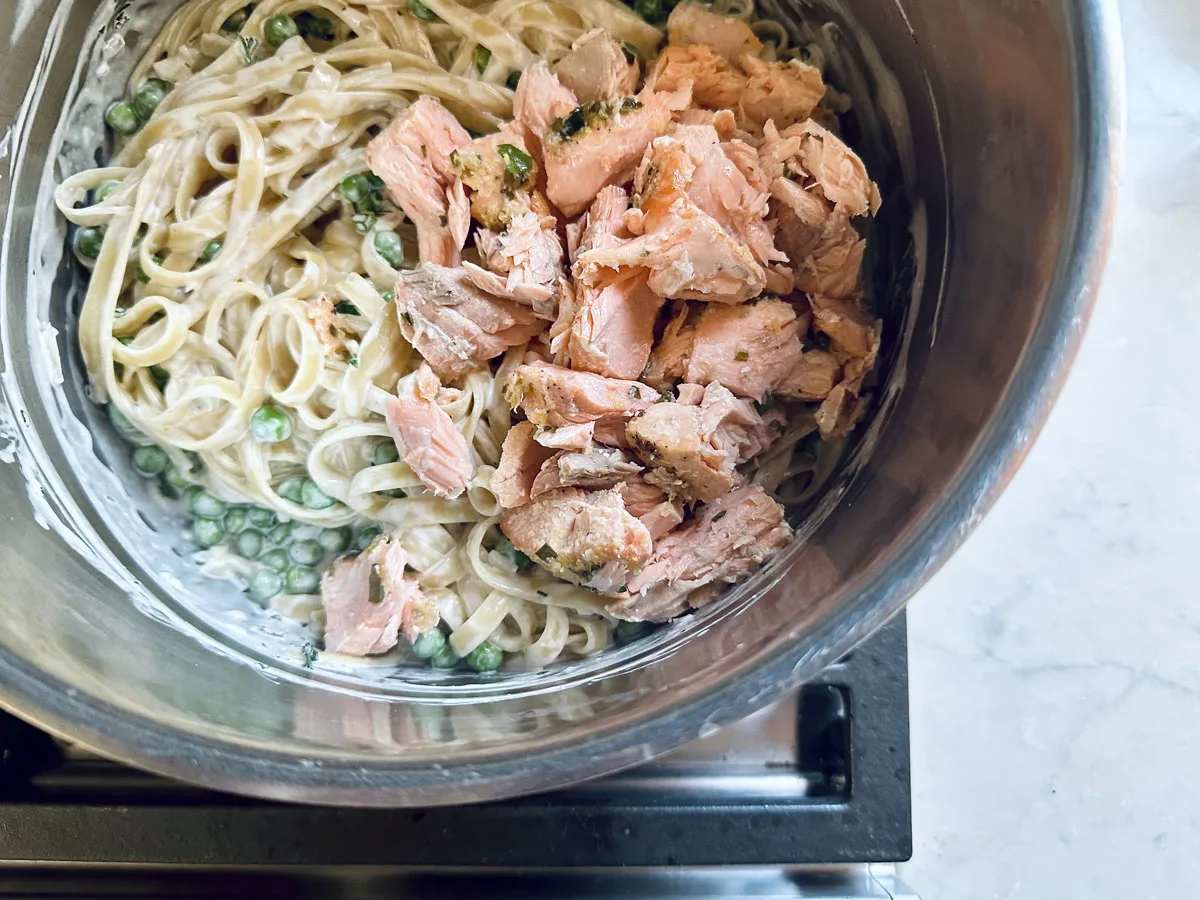 A close-up of a pot containing creamy pasta with peas and chunks of cooked salmon on top, ready to be mixed together. The pot is on a stovetop with a light-colored countertop nearby.