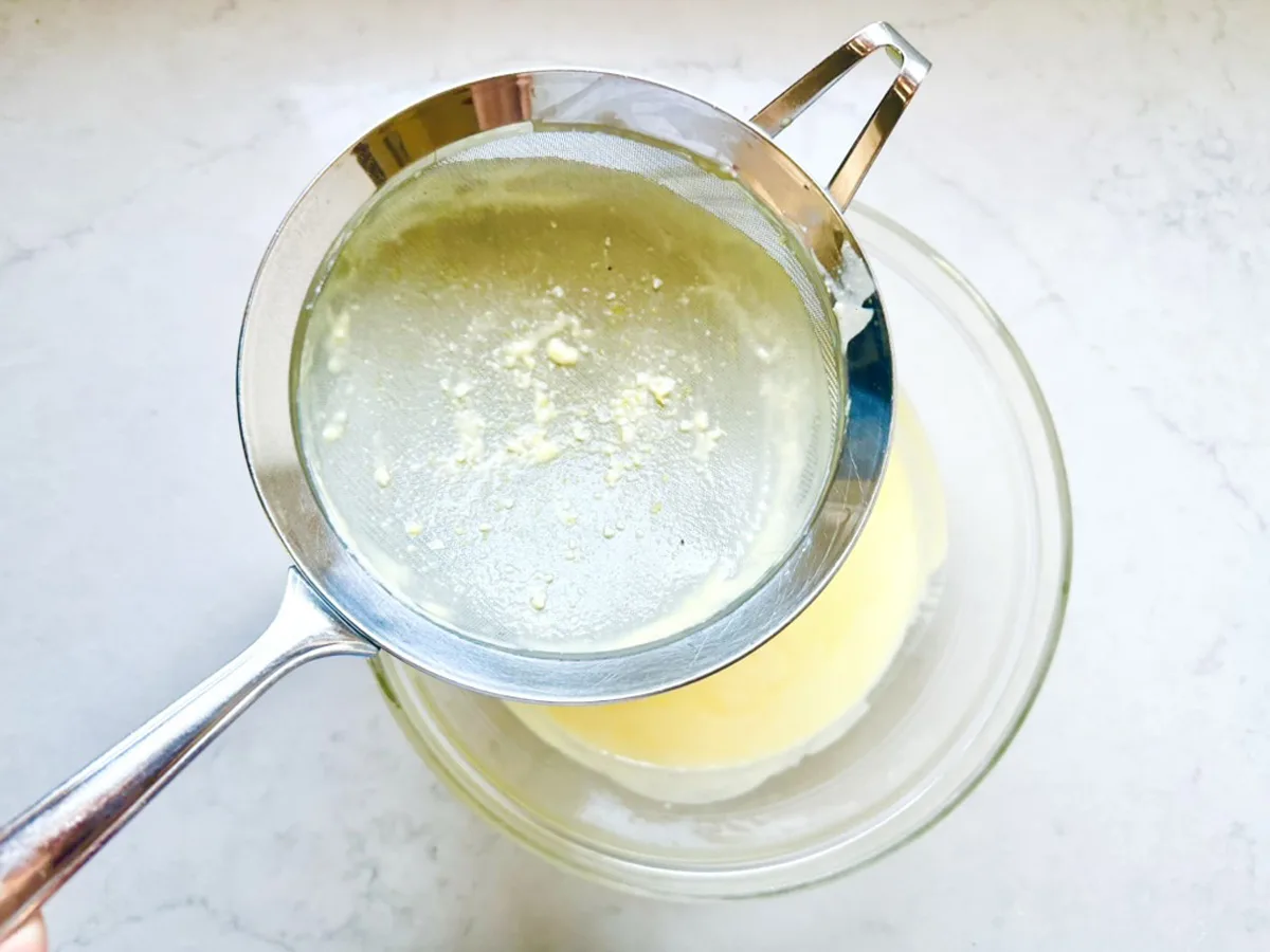 A metal strainer is held over a glass bowl, filtering a yellow liquid with small white solids separated on the strainer&mdash;perhaps prepping ingredients for Strawberry-Balsamic Ice Cream&mdash;on a light-colored countertop.