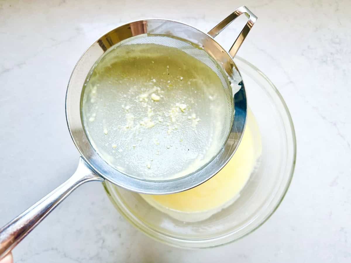 A metal strainer is held over a glass bowl, filtering a yellow liquid with small white solids separated on the strainer—perhaps prepping ingredients for Strawberry-Balsamic Ice Cream—on a light-colored countertop.