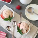 Two bowls of Strawberry-Balsamic Ice Cream, each garnished with a fresh strawberry slice and mint leaves, sit on a beige cloth. An ice cream container, scoop, extra spoons, and strawberries are nearby on a gray surface.