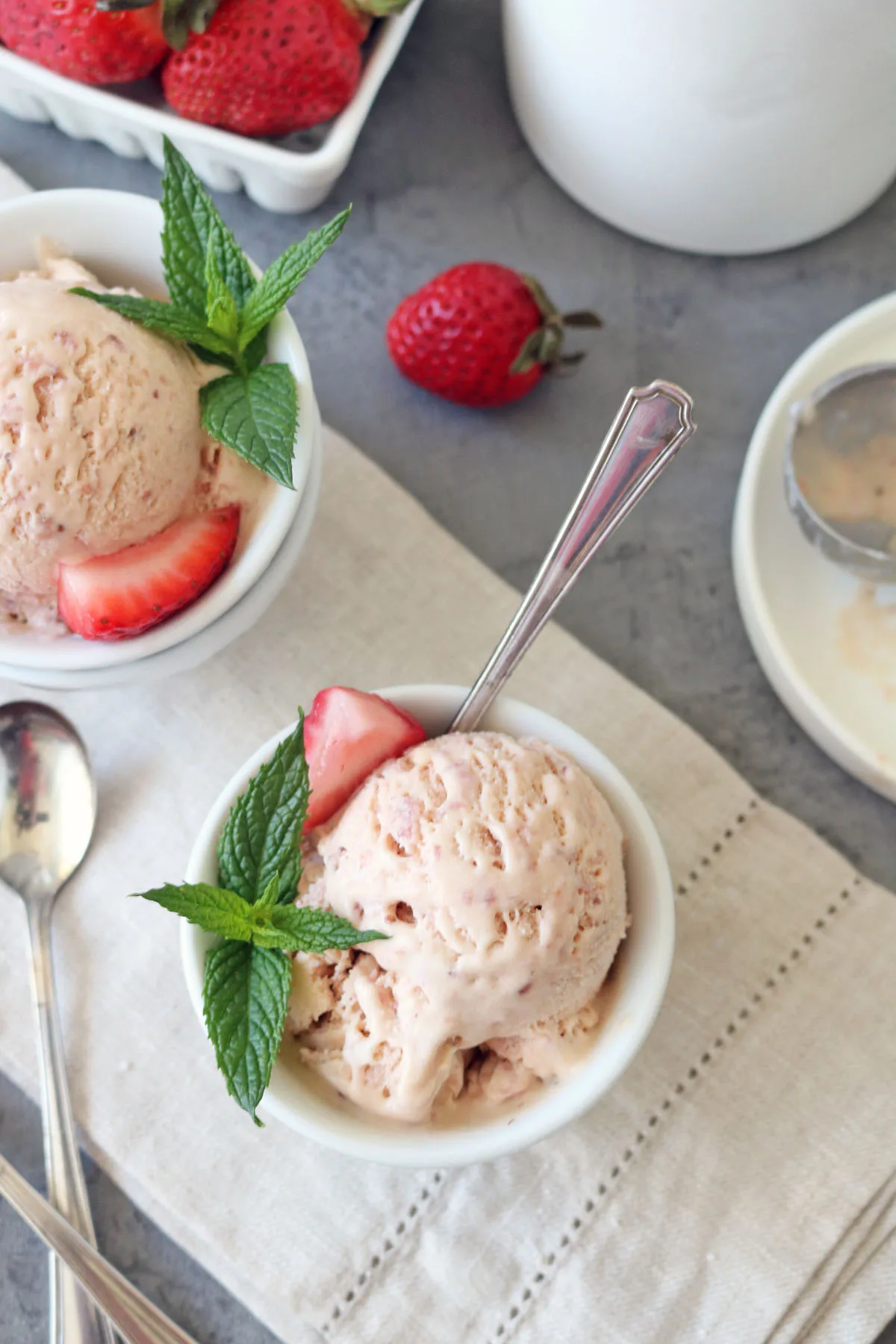 Two bowls of Strawberry-Balsamic Ice Cream garnished with fresh mint leaves and strawberry slices rest on a beige cloth, with spoons, whole strawberries, and an ice cream scoop nearby.