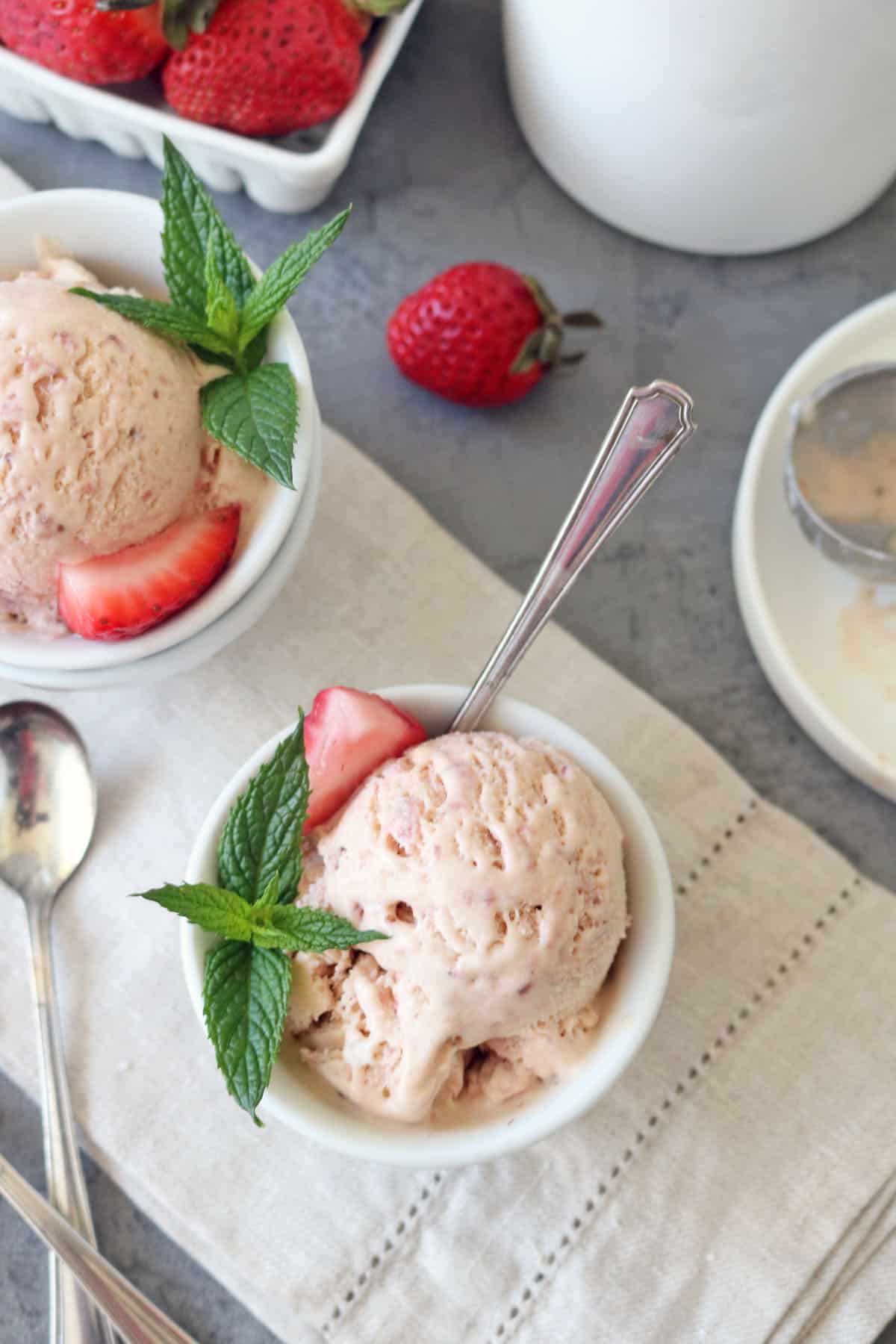 Two bowls of Strawberry-Balsamic Ice Cream garnished with fresh mint leaves and strawberry slices rest on a beige cloth, with spoons, whole strawberries, and an ice cream scoop nearby.