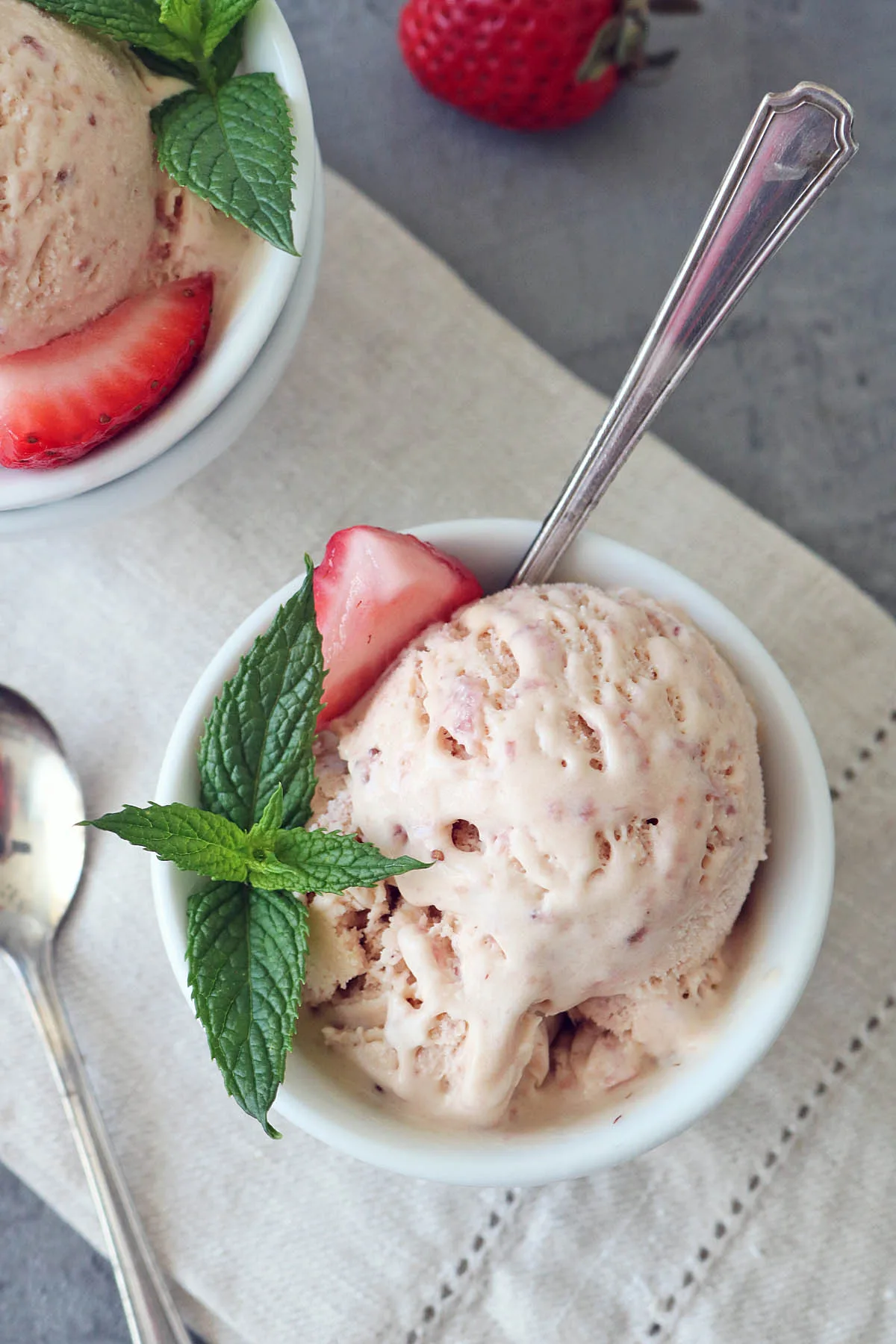 A bowl of Strawberry-Balsamic Ice Cream garnished with a fresh mint sprig and a strawberry slice, with a spoon in the bowl. Another partially visible bowl, spoon, and a whole strawberry are nearby on a light napkin.