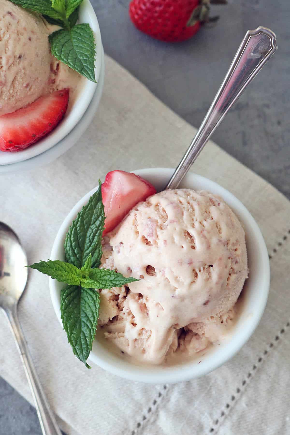 A bowl of Strawberry-Balsamic Ice Cream garnished with a fresh mint sprig and a strawberry slice, with a spoon in the bowl. Another partially visible bowl, spoon, and a whole strawberry are nearby on a light napkin.