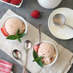 Two bowls of Strawberry-Balsamic Ice Cream, each garnished with a fresh strawberry slice and mint leaves, sit on a beige cloth. An ice cream container, scoop, extra spoons, and strawberries are nearby on a gray surface.