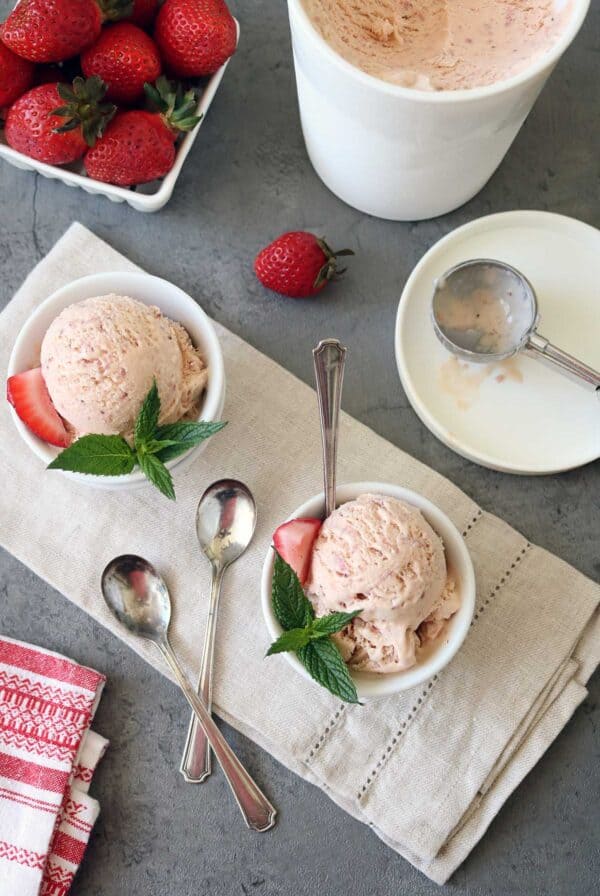 Two bowls of Strawberry-Balsamic Ice Cream, each garnished with a fresh strawberry slice and mint leaves, sit on a beige cloth. An ice cream container, scoop, extra spoons, and strawberries are nearby on a gray surface.