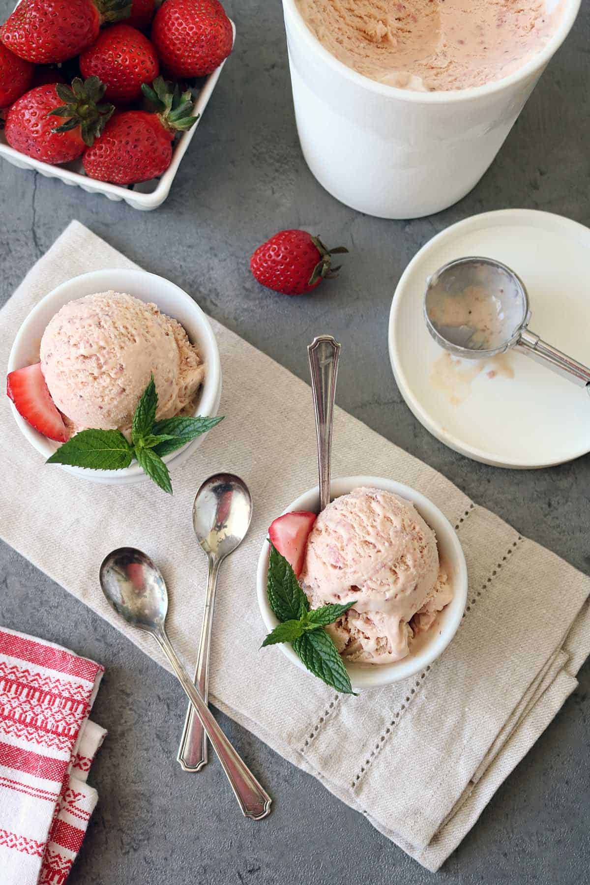 Two bowls of Strawberry-Balsamic Ice Cream, each garnished with a fresh strawberry slice and mint leaves, sit on a beige cloth. An ice cream container, scoop, extra spoons, and strawberries are nearby on a gray surface.