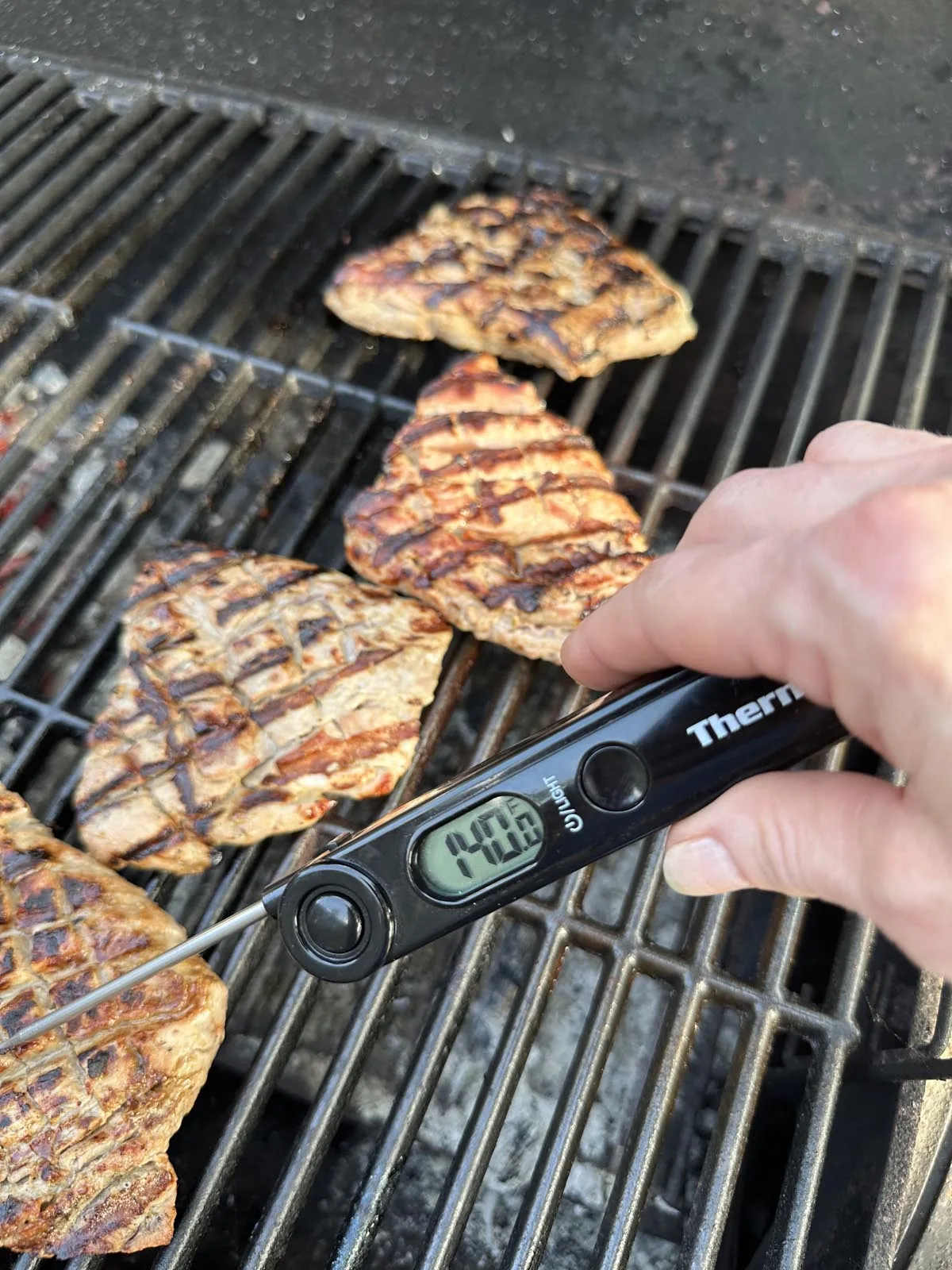 A hand holds a digital meat thermometer reading 140.8°F, checking the temperature of grilled steaks with char marks on a barbecue grill.
