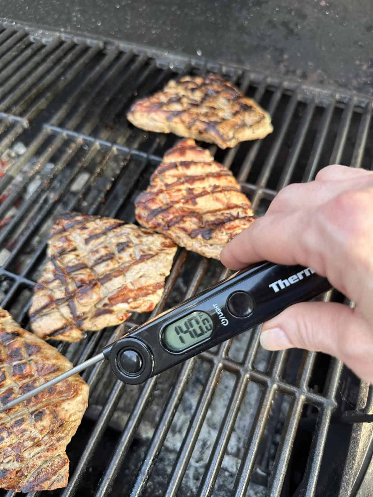 A hand holds a digital meat thermometer reading 140.8°F, checking the temperature of grilled steaks with char marks on a barbecue grill.