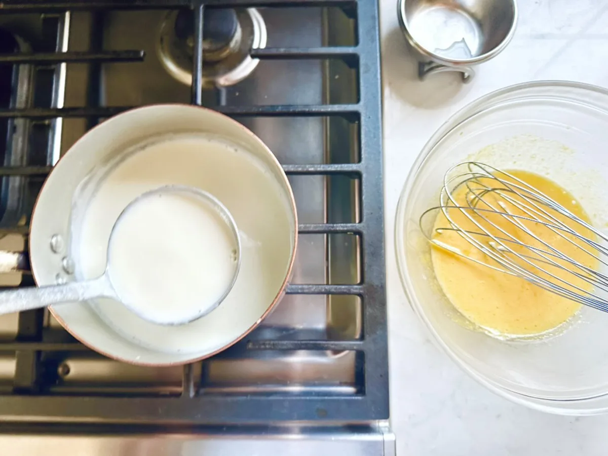 A saucepan of heated milk on a stove with a ladle inside, next to a bowl containing whisked eggs and a metal whisk&mdash;perfect prep for making creamy Strawberry-Balsamic Ice Cream on the kitchen counter.