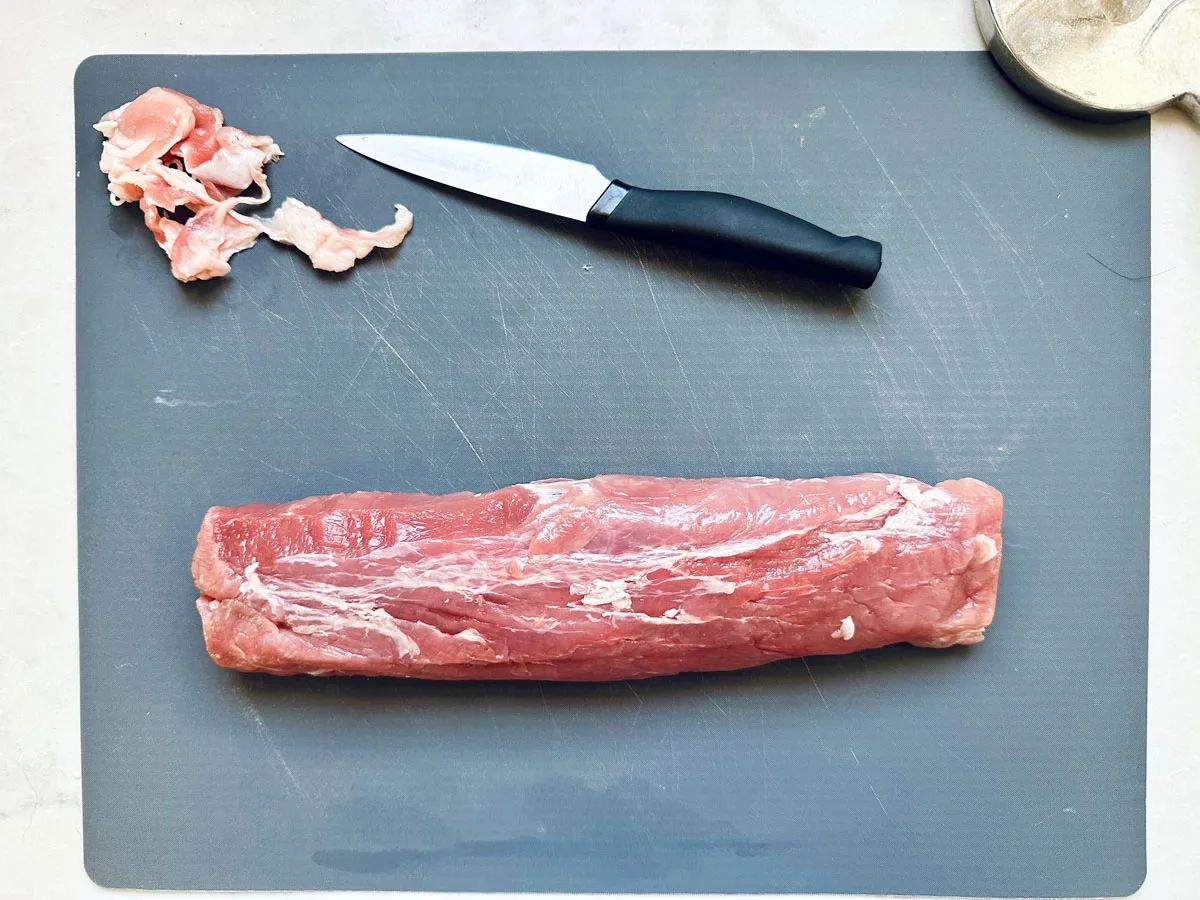 Raw pork tenderloin on a gray cutting board with a small black-handled knife above it and some trimmed fat to the side. The corner of a meat mallet is visible in the top right of the image.