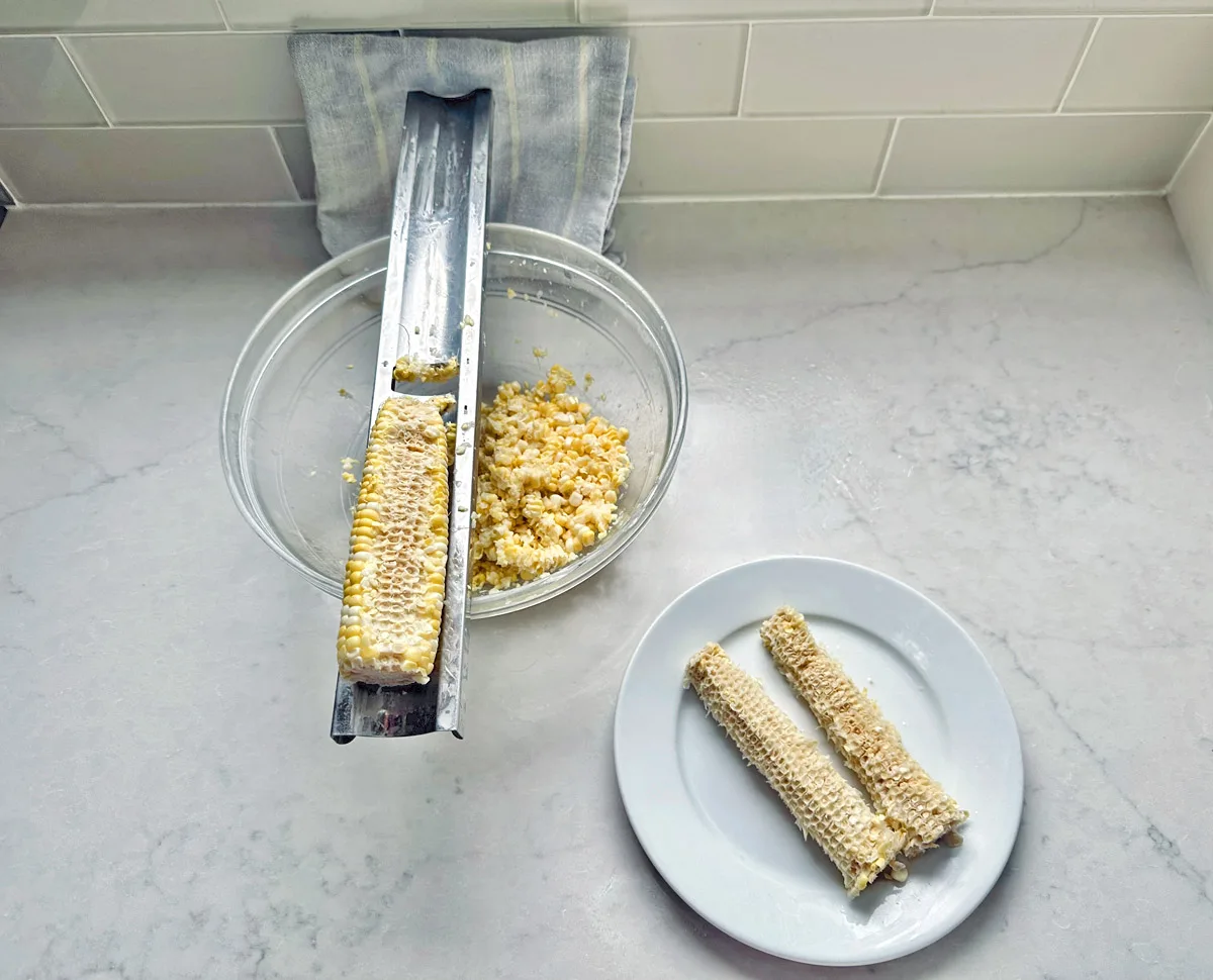 A metal corn stripper sits over a glass bowl, stripping kernels from a cob for Corn Salad with Buttermilk and Herbs. Two empty cobs rest on a white plate nearby atop a marble countertop.