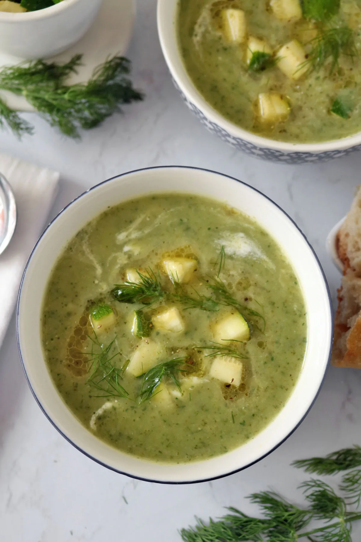 A bowl of green soup garnished with cubed vegetables and fresh dill, with a swirl of oil on top. Another bowl of soup, fresh dill, and bread are visible nearby on a light surface.