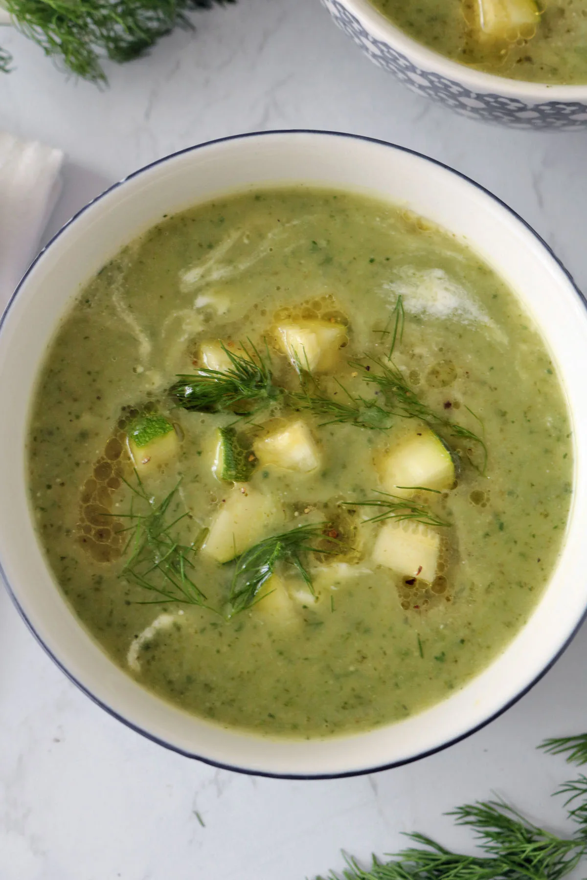 A bowl of creamy zucchini soup, garnished with diced potatoes, fresh dill, and a drizzle of olive oil, sits on a light surface with dill sprigs nearby.