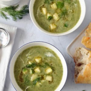 Two bowls of green soup garnished with diced zucchini and fresh herbs, placed on a white surface surrounded by bread pieces, dill sprigs, a bowl of diced zucchini, and a napkin with a spoon.