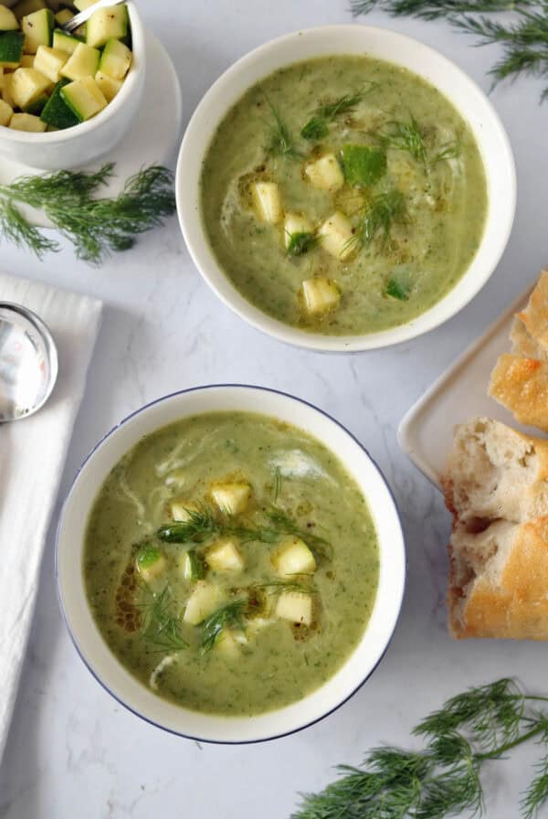 Two bowls of green soup garnished with diced zucchini and fresh herbs, placed on a white surface surrounded by bread pieces, dill sprigs, a bowl of diced zucchini, and a napkin with a spoon.