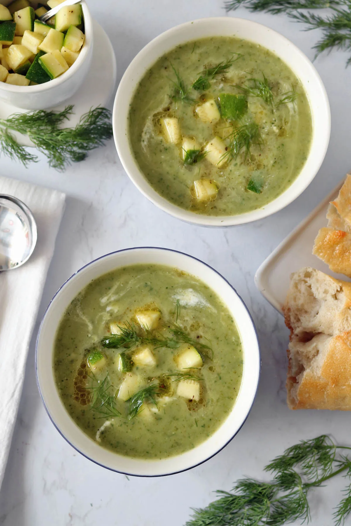 Two bowls of green soup garnished with diced zucchini and fresh herbs, placed on a white surface surrounded by bread pieces, dill sprigs, a bowl of diced zucchini, and a napkin with a spoon.