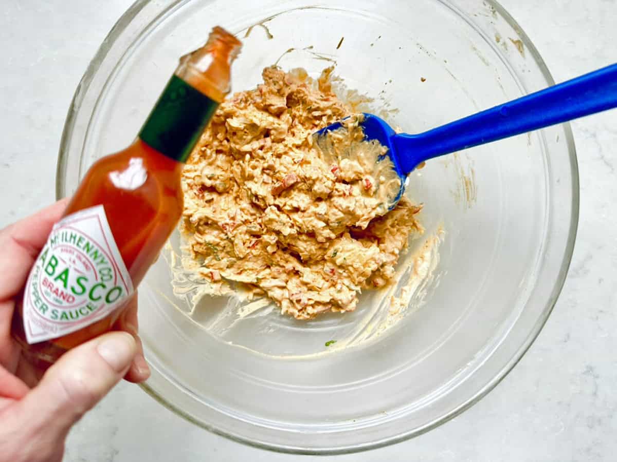 A hand holds a bottle of Tabasco sauce above a glass bowl filled with creamy Cypress Pimiento Cheese, with a blue spatula resting inside the textured mixture.