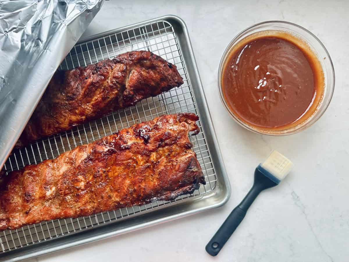 Two racks of cooked ribs on a wire rack over a baking sheet, partially covered with foil. Next to them is a bowl of barbecue sauce and a basting brush on a white countertop.