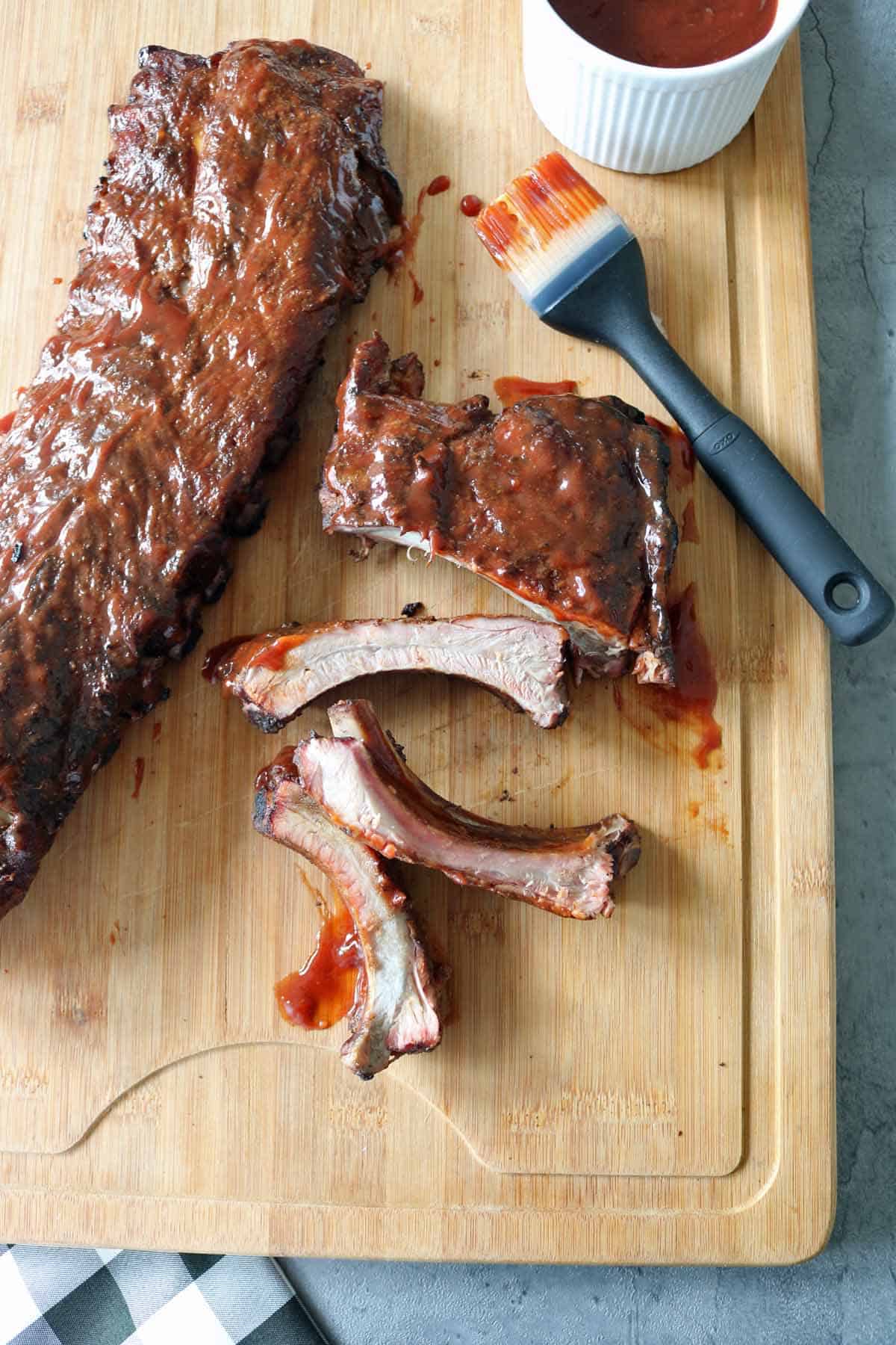 A wooden cutting board with a rack of glazed ribs, several sliced ribs, a basting brush with barbecue sauce, and a small bowl of sauce on the side. Some sauce is spread on the meat and board.