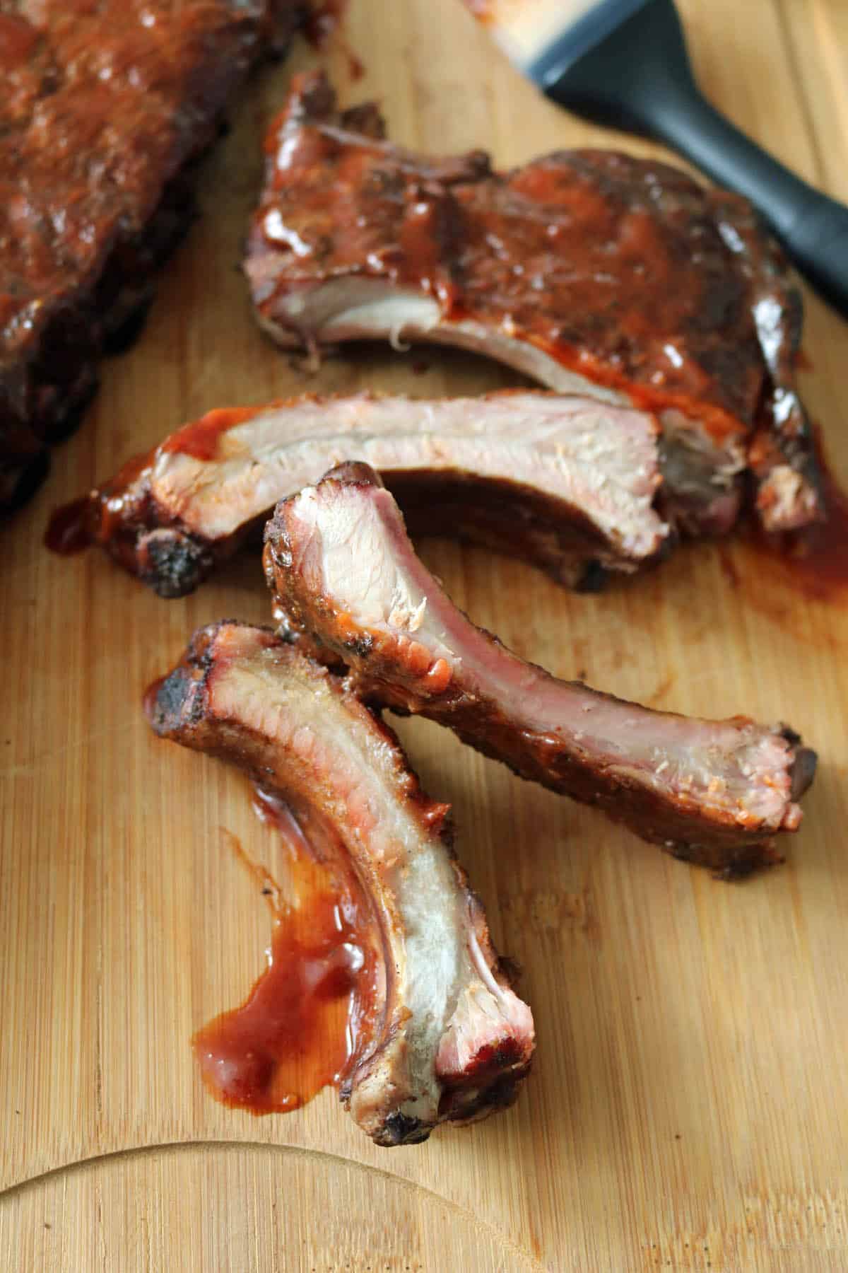 Close-up of three barbecue pork ribs with sauce on a wooden cutting board, with more ribs and a basting brush visible in the background.