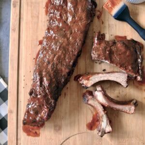 Barbecued rack of ribs on a wooden cutting board, some ribs cut and separated, with a basting brush and a small bowl of barbecue sauce nearby.