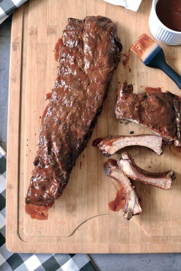 Barbecued rack of ribs on a wooden cutting board, some ribs cut and separated, with a basting brush and a small bowl of barbecue sauce nearby.