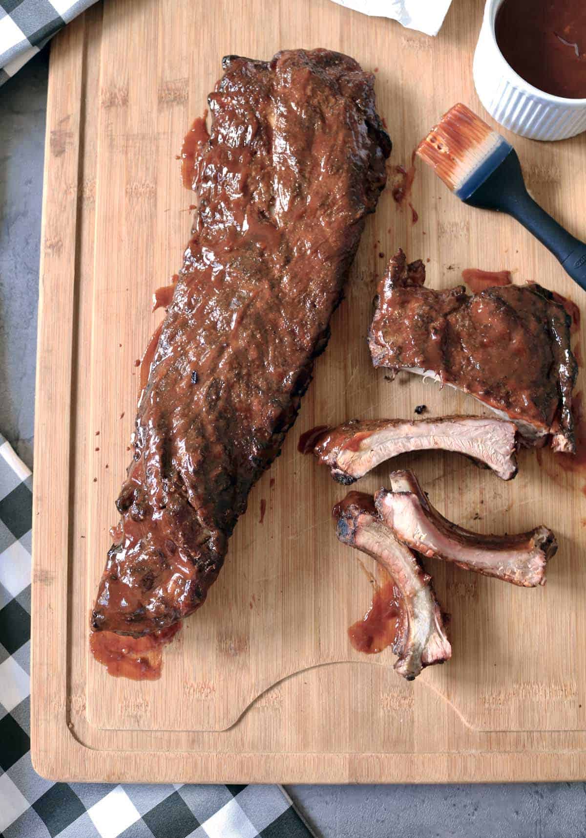 Barbecued rack of ribs on a wooden cutting board, some ribs cut and separated, with a basting brush and a small bowl of barbecue sauce nearby.