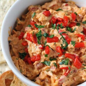 A white bowl filled with creamy, orange-hued Cypress Pimiento Cheese dip, topped with chopped red peppers and fresh herbs, surrounded by toasted bread slices and crackers on a plate.