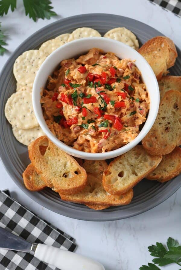 A bowl of creamy Cypress Pimiento Cheese dip garnished with chopped herbs and red peppers is surrounded by toasted baguette slices and round crackers on a gray plate, all set on a marble surface with a checkered napkin and parsley leaves nearby.