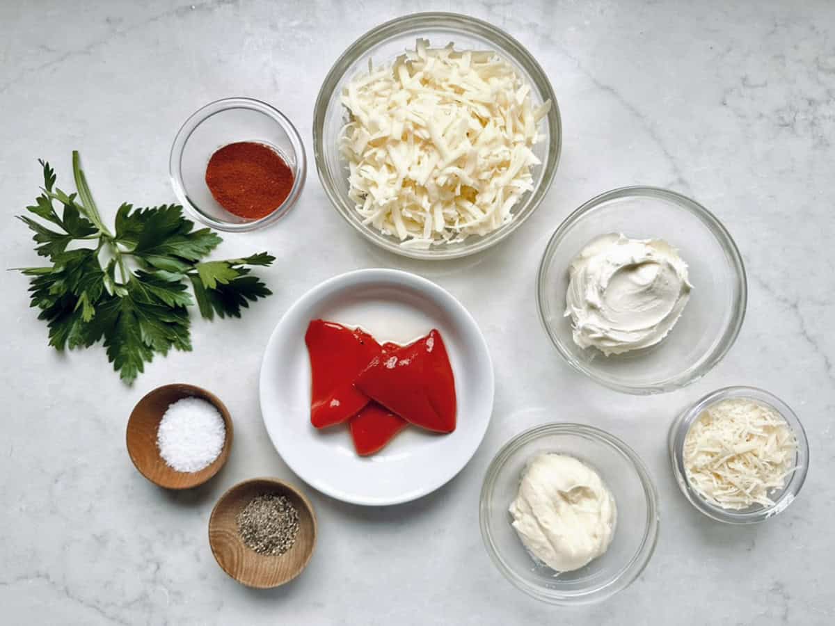 A flat lay of ingredients for Cypress Pimiento Cheese in bowls on a white surface: shredded cheese, cream cheese, mayonnaise, grated Parmesan, red peppers, parsley, paprika, salt, and pepper.