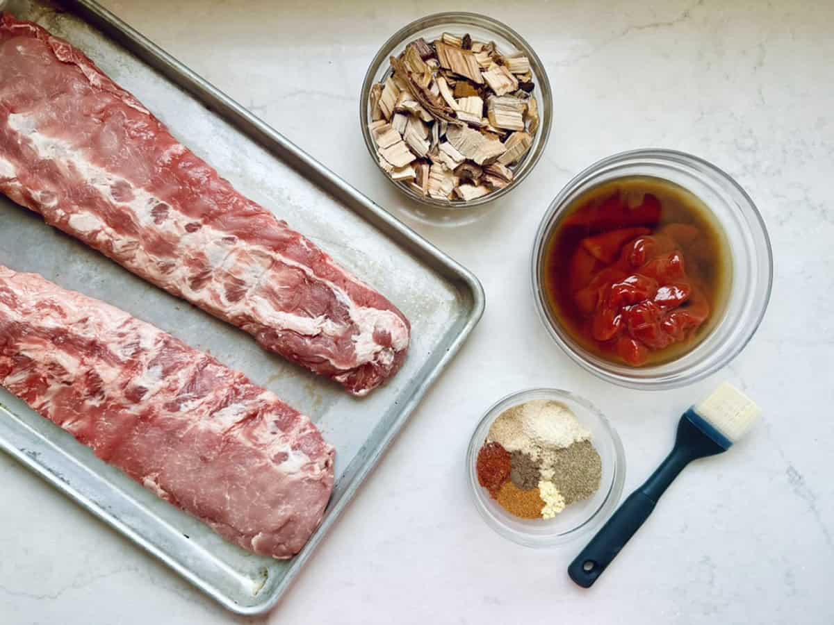 Two raw pork ribs on a metal tray next to bowls of wood chips, barbecue sauce with chopped tomatoes, assorted spices, and a basting brush on a white countertop.