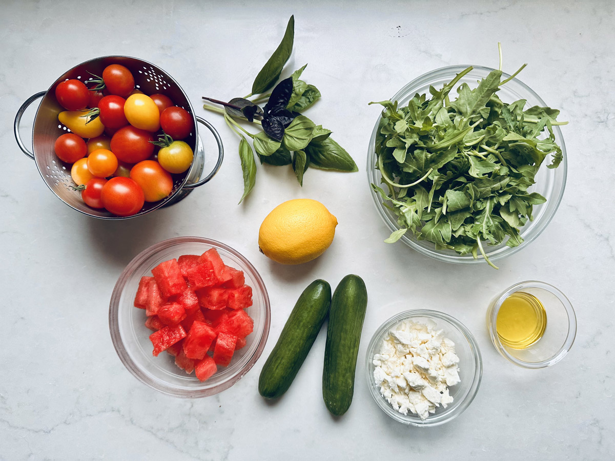 A colander of tomatoes, fresh basil, a bowl of arugula, a lemon, cubed watermelon, two cucumbers, crumbled feta cheese, and a small glass of olive oil on a white surface—everything you need to prepare a refreshing Tomato and Watermelon Salad with Feta Cheese.