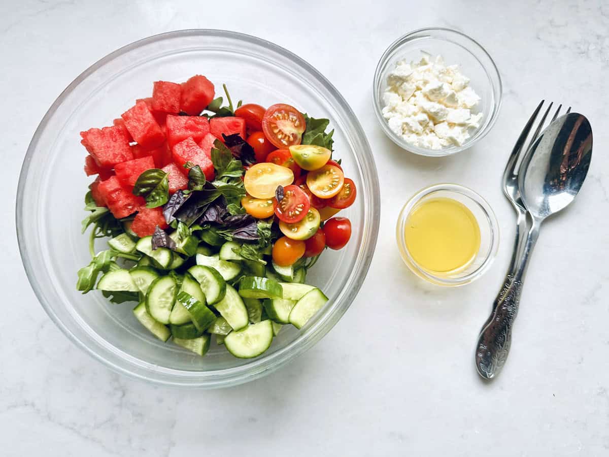 A glass bowl with Tomato and Watermelon Salad with Feta Cheese, featuring juicy watermelon, ripe tomatoes, crisp cucumbers, and fresh greens. Next to it are small bowls of feta cheese and dressing, plus a spoon and fork on a white surface.