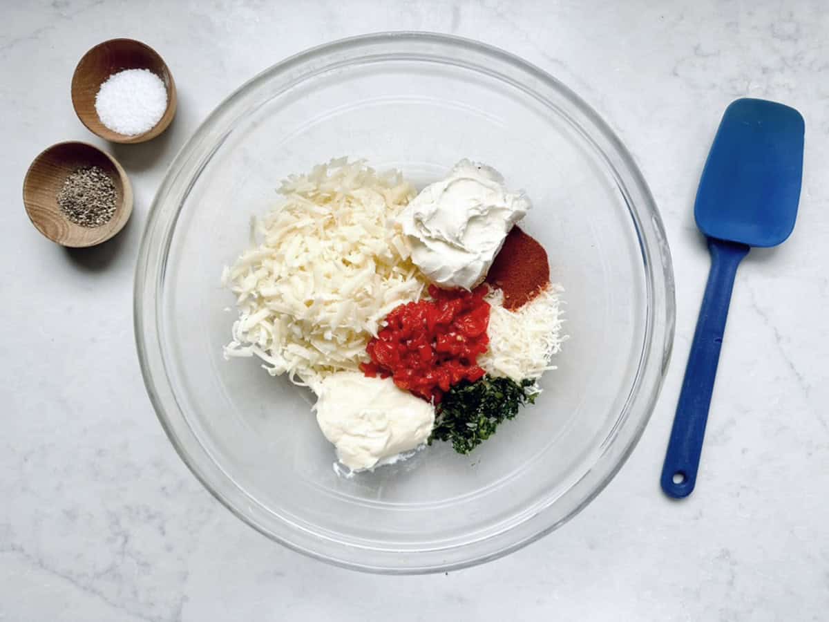 A glass bowl contains Cypress Pimiento Cheese made with shredded cheese, cream cheese, mayonnaise, red diced peppers, chopped herbs, and spice. Nearby are a blue spatula and small wooden bowls with salt and pepper on a white countertop.