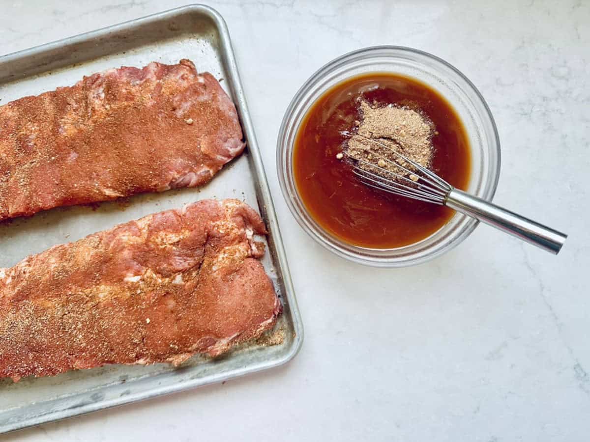 Two racks of uncooked, seasoned ribs are on a metal baking tray next to a glass bowl of sauce being mixed with a whisk on a white countertop.
