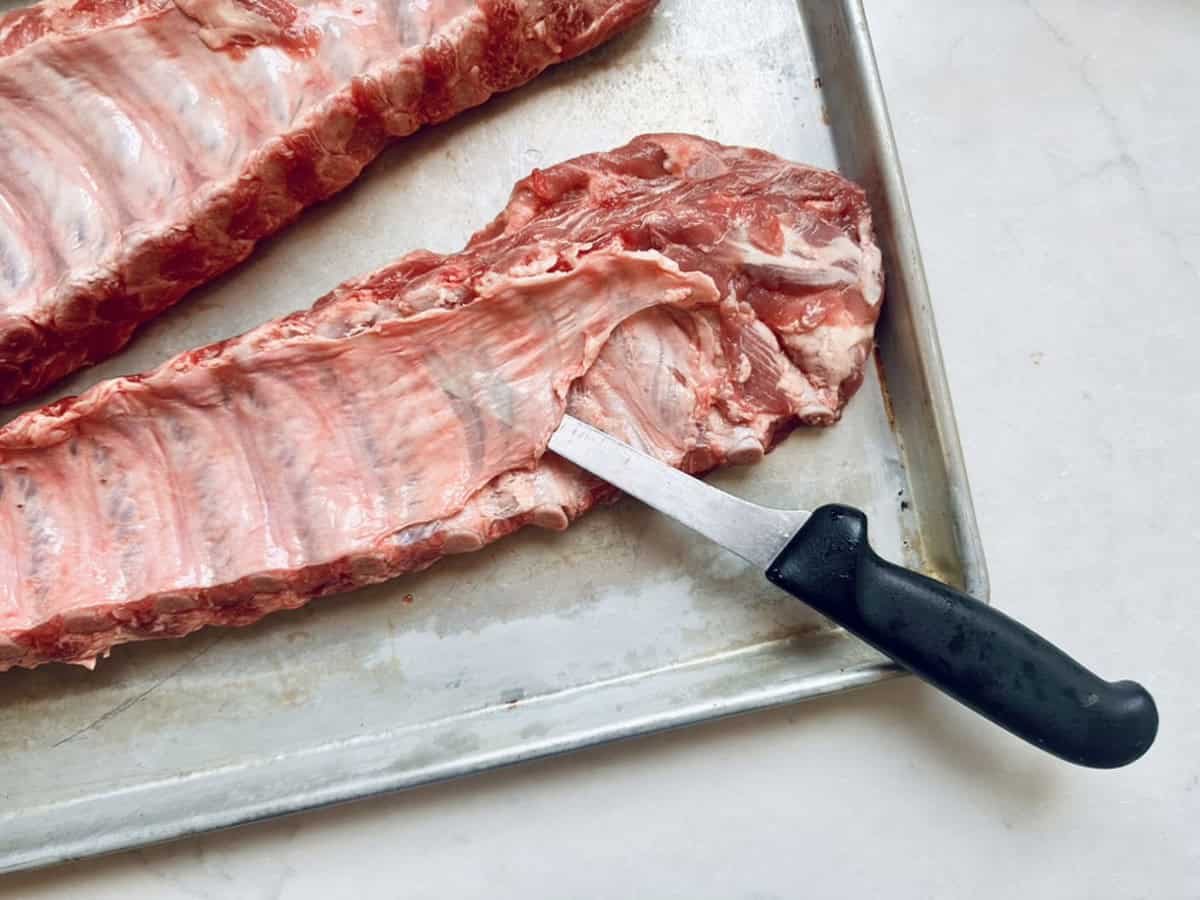 A close-up of a raw rack of ribs on a metal tray, with a knife inserted under the membrane to separate it from the meat.