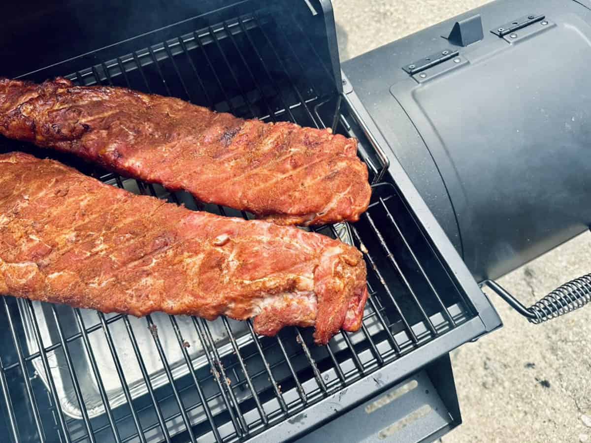 Two racks of seasoned ribs cooking on the grill of a smoker, with smoke visible rising around the food and the smoker lid partially open.
