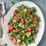 A white oval plate holds a fresh Tomato and Watermelon Salad with Feta Cheese, featuring arugula, cucumber, cherry tomatoes, olives, and juicy watermelon. A spoon and napkin are on the side, with a small bowl of salad and basil leaves nearby.