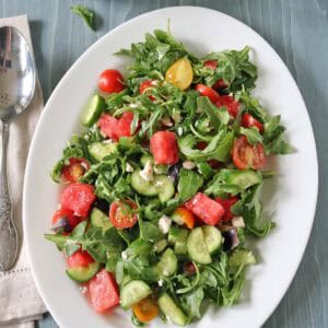 A white oval plate holds a fresh Tomato and Watermelon Salad with Feta Cheese, featuring arugula, cucumber, cherry tomatoes, olives, and juicy watermelon. A spoon and napkin are on the side, with a small bowl of salad and basil leaves nearby.