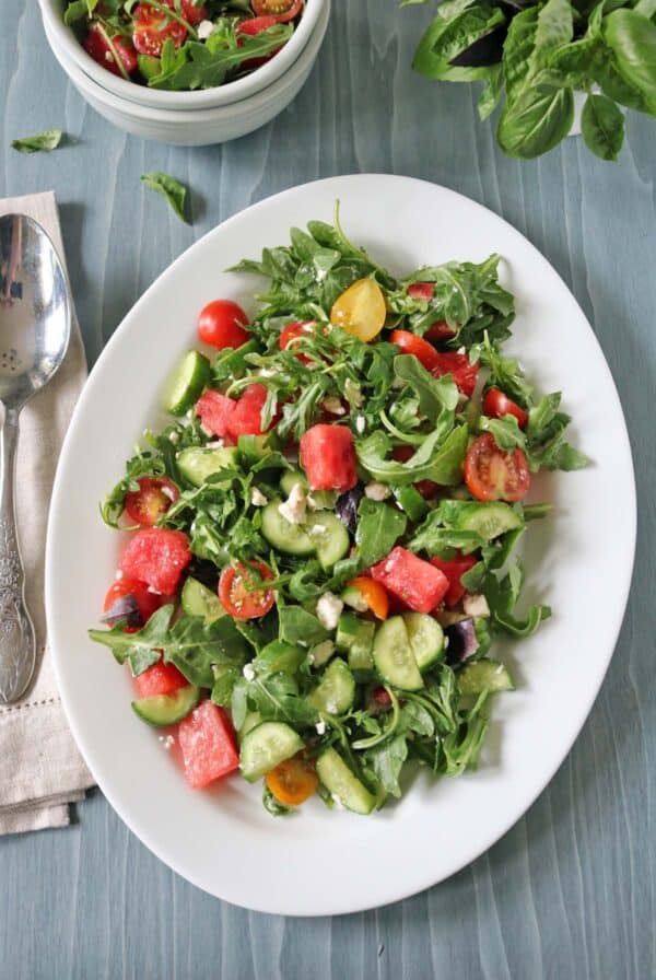 A white oval plate holds a fresh Tomato and Watermelon Salad with Feta Cheese, featuring arugula, cucumber, cherry tomatoes, olives, and juicy watermelon. A spoon and napkin are on the side, with a small bowl of salad and basil leaves nearby.
