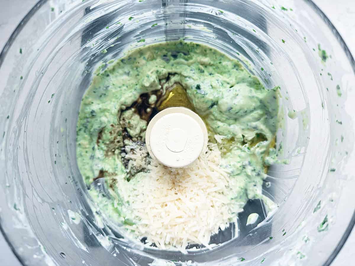 A food processor bowl contains a creamy green mixture, likely pesto, with grated cheese, olive oil, and herbs visible on top, ready to be blended.