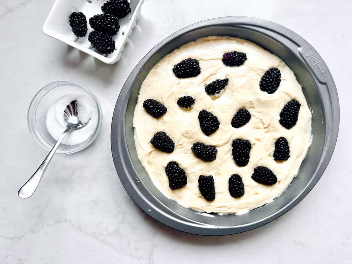 A round baking pan with cake batter topped with fresh blackberries, next to a small bowl of sugar with a spoon and a dish of extra blackberries on a white surface.
