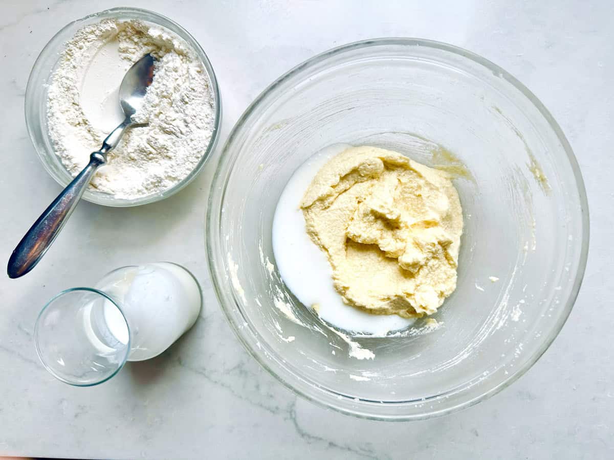 A glass bowl with yellow batter and milk, a bowl of flour with a spoon, and a small glass of white liquid sit on a white countertop.