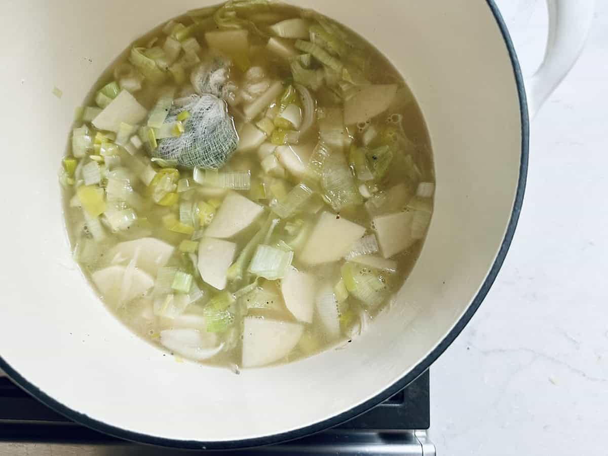 A white pot on a stove contains Potato Leek Soup, with chopped leeks, potatoes, broth, and a wrapped bundle of herbs gently simmering.